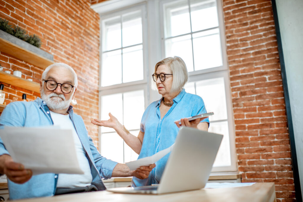 Senior couple with shocked emotions looking on high bills or taxes sitting with laptop on the kitchen at home